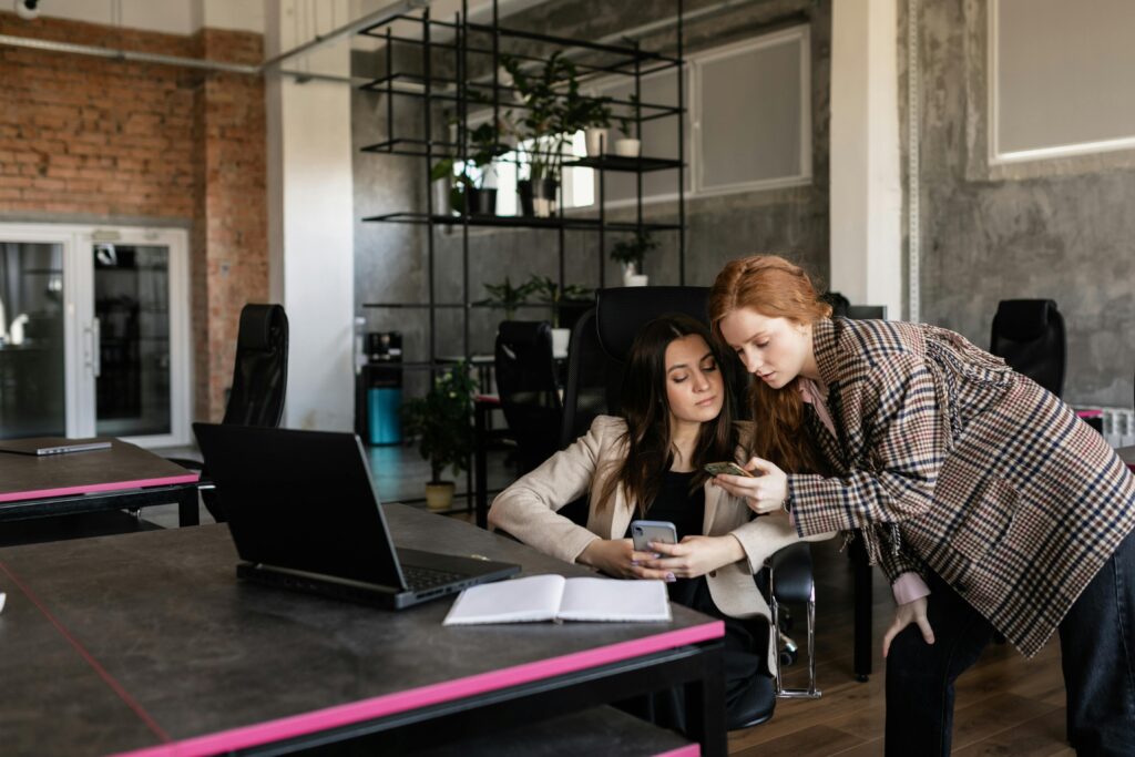 Two women collaborating with a smartphone in a modern open office setting. Teamwork and technology.