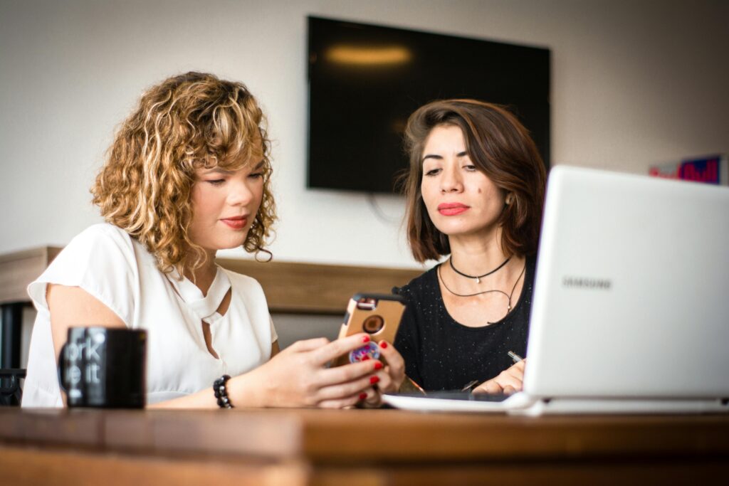 Two women using a smartphone and laptop in a modern office setting, sharing ideas.